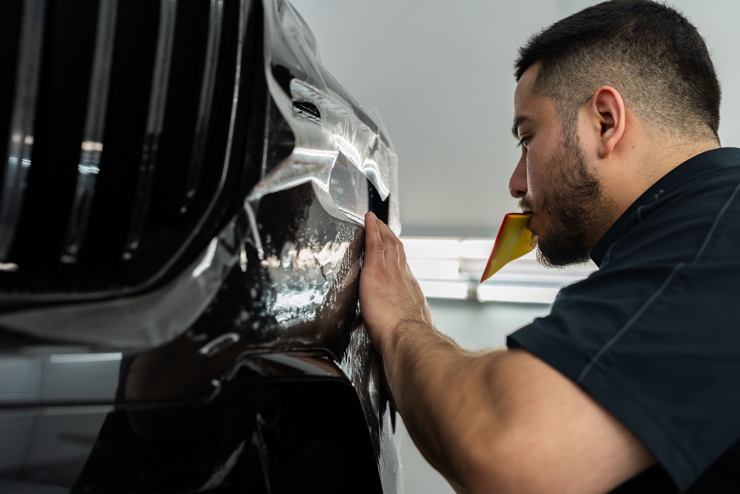 Man perfects clear PPF placement on vehicle hood with squeegee in mouth